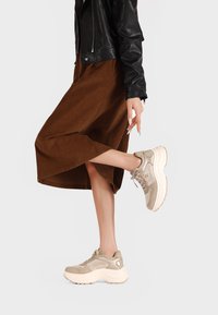 Sneaker beige con una robusta suola bianca, superiore in suede testurizzato e mesh, abbinate a una gonna marrone di lunghezza media e a una giacca di pelle nera.