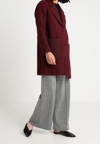 Burgunderfarbener, strukturierter Mantel mit langen Ärmeln und Fronttaschen, kombiniert mit grauen Hahnentrittmuster-Hosen in weit geschnittener Form und schwarzen spitzen Slippern.