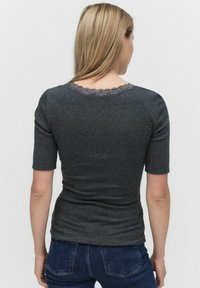 Kvinde med blondt hår iført en mørkegrå kortærmet bluse med blonder i halsudskæringen, parret med mørkeblå jeans, stående foran en ensfarvet baggrund.