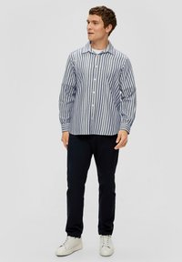 Gestreepte lange mouwen shirt in marineblauw en wit, met knoopsluiting en zak; gedragen met marineblauwe broek en witte sneakers op een neutrale achtergrond.