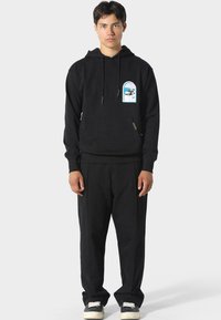 Joven vestido con una sudadera negra con un gráfico de coche y pantalones negros a juego, de pie frente a un fondo claro y uniforme.