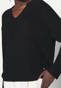 Svart ribbad stickad tröja med V-hals, långa ärmar och sprund på sidorna. Har en texturerad yta och subtil logotypdetalj vid fållen.