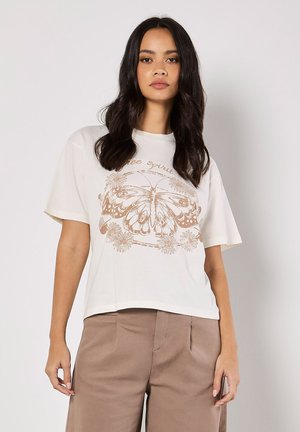 T-shirt en coton couleur crème avec des manches courtes, présentant un graphique de papillon marron et des éléments floraux imprimés sur le devant.