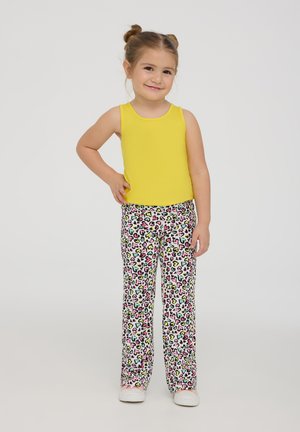 Top giallo senza maniche abbinato a pantaloni bianchi con un motivo a cuori multicolori e stampa leopardata. Il bambino sorride e posa con le mani sui fianchi.