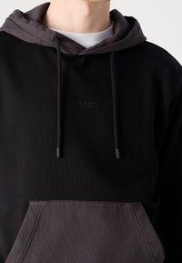 Sudadera negra con capucha y bolsillo en gris contrastante. Presenta un branding minimalista con un sutil logo bordado de "BOSS" en el pecho.
