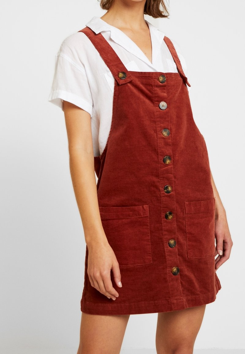 Femme portant une robe salopette en velours côtelé couleur rouille avec des boutons sur le devant, par-dessus une chemise blanche à manches courtes et col, debout devant un fond uni.