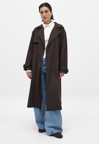 Brun lång trenchcoat med bred krage, dubbelknäppning och vida ärmar, parat med blå vida jeans och mörka skor.