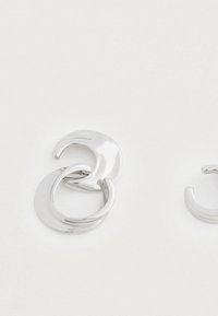 Masívne lesklé strieborné náušnice v tvare dvojitých kruhov s nepravidelnými zaoblenými tvarmi na jednoduchom bielom pozadí.