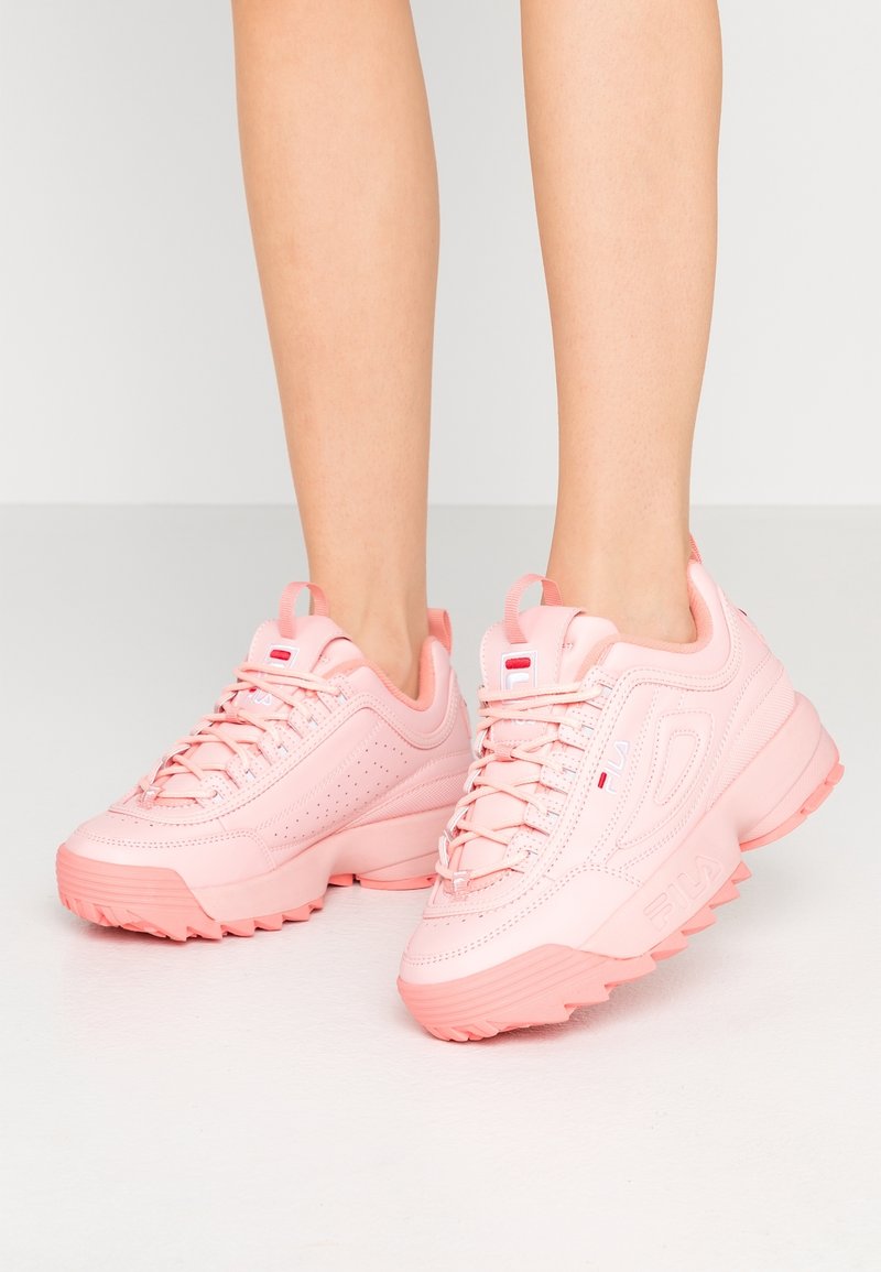 Jambes portant des baskets roses épaisses avec des semelles épaisses, des coutures détaillées et des lacets, debout sur un sol clair uni.