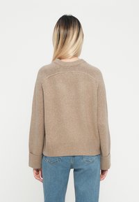 Beige pletený sveter s okrúhlym výstrihom, rebrovanými manžetami a lemom. Má voľný strih a jemnú textúru, nosí sa cez modré džínsy.