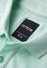 Polo shirt verde menta in tessuto texturizzato; presenta un'etichetta nera con la scritta "ombre" e dettagli sulla taglia cuciti nel colletto.