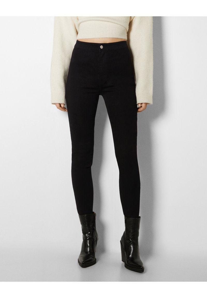 Jeans noirs taille haute en tissu extensible, avec une coupe slim et une fermeture à boutons sur le devant. Assortis avec des bottines noires texturées.