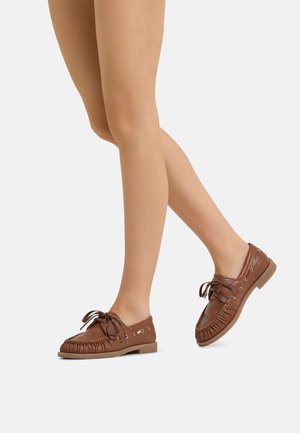 Bare legs wearing brown leather moccasin loafers with lace-up details, shown walking against a plain light background.