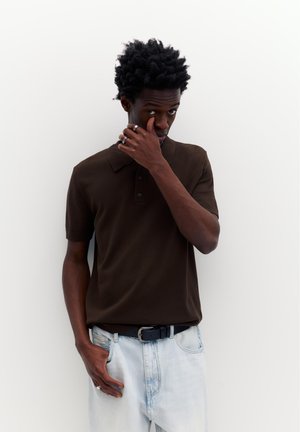 Giovane con capelli mossi che indossa una polo marrone scuro e jeans azzurri, si sistema il viso con una mano, in piedi contro un muro bianco.