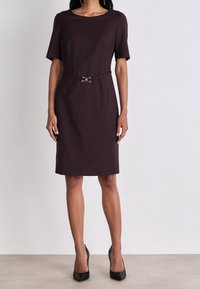 Burgundské šaty ke kolenům s krátkým rukávem, kulatým výstřihem a páskem v pase s detaily ve tvaru stříbrné spony. Hladká textilie.