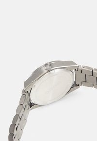 Montre en acier inoxydable argenté avec un boîtier rond, couronne discrète gravée "HUGO" et un bracelet à maillons. Présente un design minimaliste.