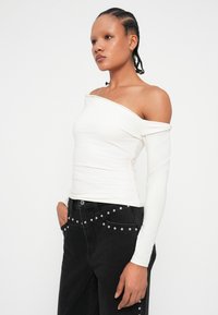 Bela off-shoulder bluzica z dolgimi rokavi, prilegajoča zasnova. V kombinaciji z visokim pasom črnimi kavbojkami, ki imajo srebrne čepke ob pasu.