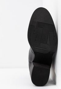 Chaussure en cuir noir avec une semelle en caoutchouc texturée dotée d'une accroche à motif ; vue en angle montrant les détails du talon et de la semelle extérieure.
