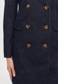 Granatowy, dwurzędowy płaszcz denimowy o teksturowanym wykończeniu, z sześcioma okrągłymi brązowymi guzikami i delikatnym kontrastowym szwem na kieszeniach.