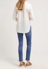 Žena stojící zády, oblečená v bílé blůze s dlouhým rukávem, modrých slim-fit džínsech a béžových volnočasových teniskách na neutrálním pozadí.