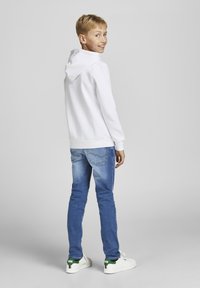 Felpa bianca con cappuccio e cordoncino, jeans blu e sneakers bianche con dettagli verde. Il bambino è visto di spalle, sorridendo.