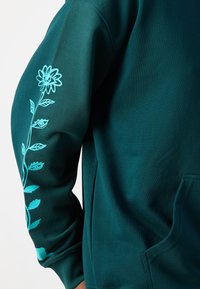 Hoodie teal scuro con un design floreale in blu chiaro sulla manica sinistra. Il tessuto sembra morbido e presenta una tasca sulla parte frontale.