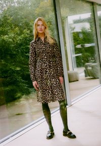 Suknja z leopardjim vzorcem, s kapuco, dolgimi rokavi in dolžino do kolen, v kombinaciji s temnimi vzorčenimi nogavicami in črnimi loaferji.