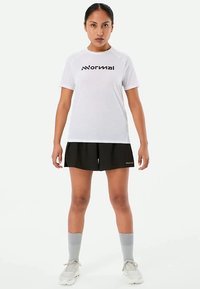 Baltas trumpas rankovėmis sportinis marškinėlis su juodu logotipu, derinamas su juodomis šortais ir pilkomis iki kelių kojinėmis, stovintis ant vienodo fono.