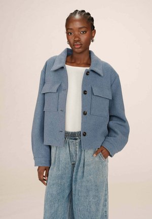 Veste en laine bleue coupée avec poches avant, grand col et boutons noirs, associée à un pantalon en denim bleu clair taille haute.