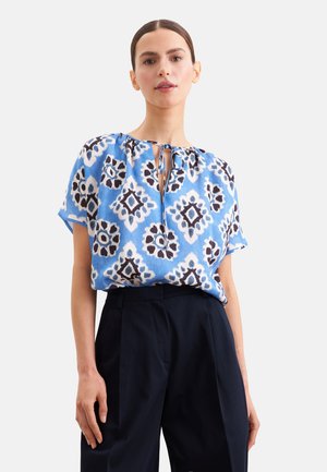 Vrouw draagt een blauw gemêleerde korte mouwen blouse met een striksluiting bij de hals en een zwarte broek met hoge taille, staand tegen een egale achtergrond.