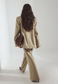 Bežirano krojen jopič z dolgim blazerjem in širokimi hlačami. Oseba drži rjav pokrovček. Mehak material in gladka tekstura.