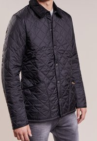 Homme portant une veste noir matelassée avec un col texturé, fermeture à boutons sur le devant et poches pour les mains, associée à un jean gris.