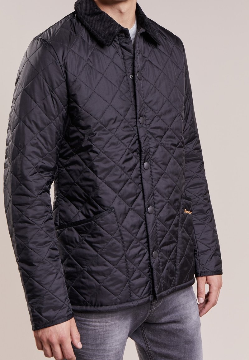 Homme portant une veste noir matelassée avec un col texturé, fermeture à boutons sur le devant et poches pour les mains, associée à un jean gris.