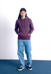 Sudadera morada con bolsillo frontal y cordones, combinada con jeans azul claros y zapatillas negras y blancas, de pie sobre una alfombra azul.