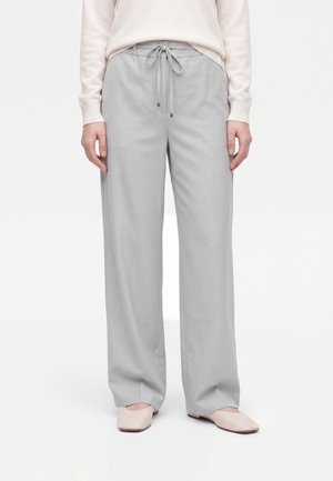 Personne portant un pantalon ample gris clair avec une taille à cordon, un haut crème et des chaussures plates beiges, debout sur un fond blanc.