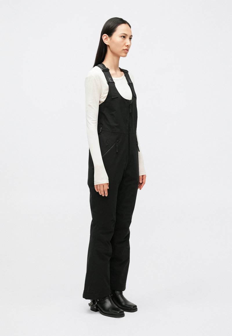 Black overalls with adjustable straps and zippered pockets, paired with a long-sleeve cream top. Smooth fabric and fitted design.