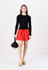 Čierny dlhý kardigán s gombíkmi, skombinovaný s červenou plisovanou sukňou, čiernou vzorovanou kabelkou a béžovými platformovými sandálmi.