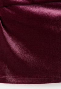 Burgundský samet s hladkou strukturou, jemným leskem a zaobleným lemem. Švy jsou viditelné na spodním okraji.