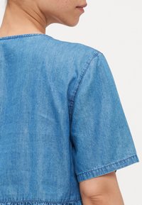 Denimska srajca s kratkimi rokavi in zaobljenim izrezom, izdelana iz modre tkanine z diskretno teksturo. Šivi so vidni ob šivih.