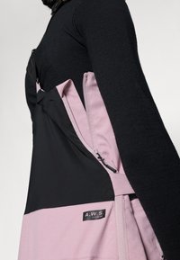Svart og rosa yttertøy med glidelåsåpning på siden, laget av mykt stoff, med en tydelig "A.W.S. EXTREME"-lapp.