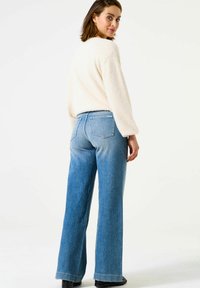 Jeans larges bleu clair à taille haute, dotés de poches arrière et d'un léger dégradé. Portés avec un pull texturé crème.