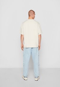 T-shirt oversized beige clair à manches courtes, associé à un pantalon cargo bleu clair avec des poches latérales et des poignets élastiques, porté avec des baskets.