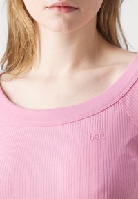 Femme portant un haut rose côtelé de la marque Lee avec un large décolleté dévoilant son cou, ses clavicules et le bas de son visage.