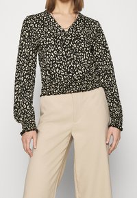 Mustikka- ja beige leopardikuosiset puserot, joissa on V-pääntie, pitkät pallohihat ja rypytetty helma, yhdistettynä vaaleanbeigeihin leveälahkeisiin housuihin.