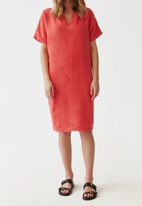 Kleid aus Korallenleinen mit kurzen Ärmeln, V-Ausschnitt, geradem Schnitt und Nahtdetails auf der Vorderseite. Schwarze Sandalen mit Schnallenakzenten.