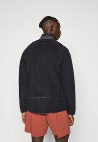 Schwarze Fleecejacke mit hohem Kragen und strukturiertem Stoff, mit kontrastierenden Nähten und lässiger Passform, kombiniert mit orangen Shorts.