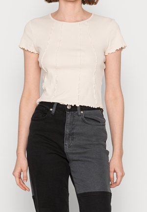 Vrouw met een crème ribgebreide top met korte mouwen en een golvende zoom, gecombineerd met zwart-grijze high-waisted jeans met twee tinten.