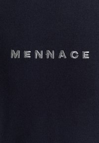 Tissu foncé avec le mot "MENNACE" brodé en fil gris, uniformément espacé au centre.