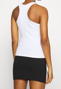 Kvinna iförd en vit racerback linne och en svart minikjol, visad bakifrån mot en enkel vit bakgrund.