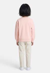 Παιδί με κοντά μαύρα μαλλιά, που στέκεται πλάτη, φοράει ροζ ανοιχτό φούτερ, μπεζ παντελόνι και λευκά αθλητικά παπούτσια σε λευκό φόντο.